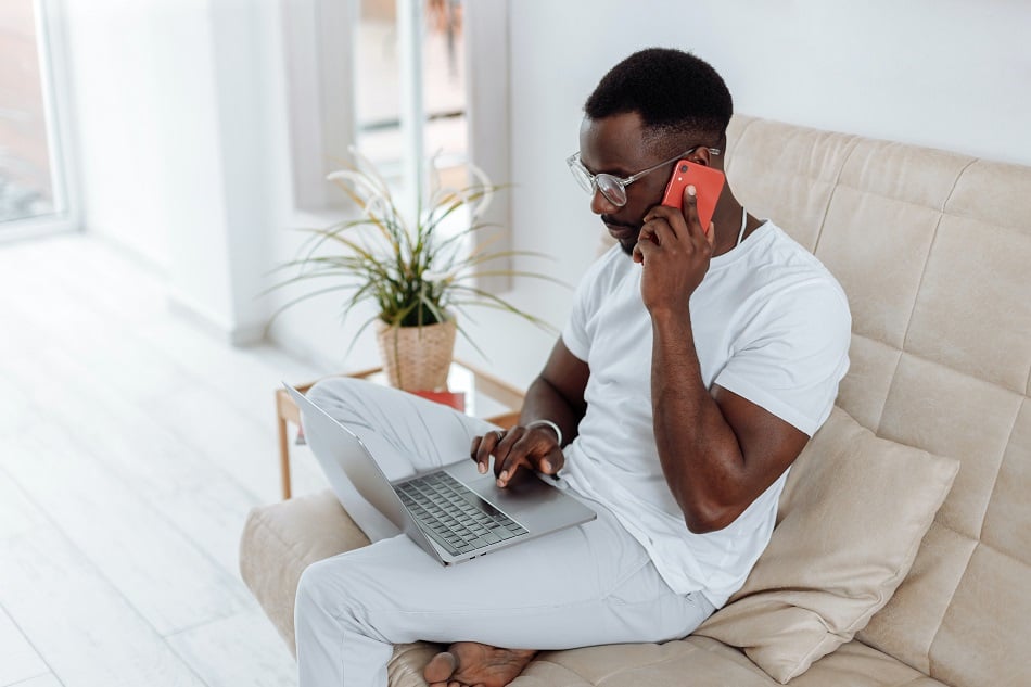 Man sits working on his laptop and phone, from his sofa 